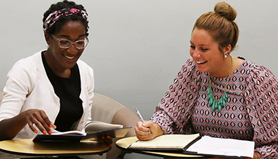 Two students talking with each other while taking notes from books.